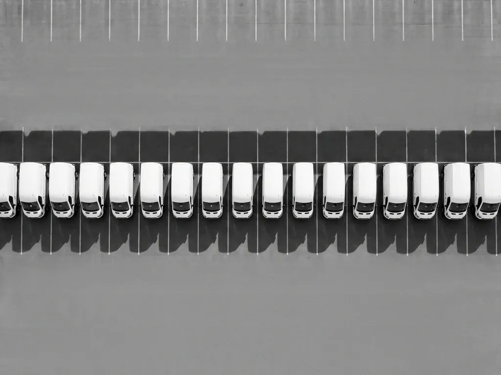 Witte bedrijfsvoertuigen in perfecte rij op grijze parkeerplaats, bovenaanzicht met geometrische schaduwen