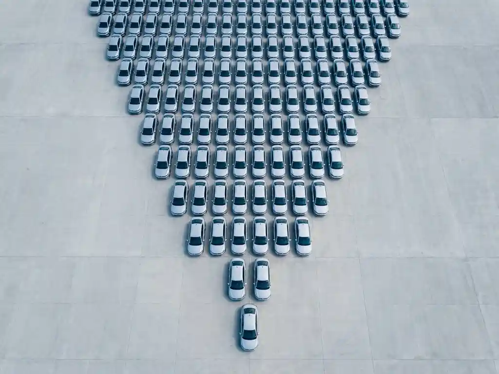 Luchtfoto van een groeiende vloot identieke zilveren sedans in driehoeksformatie op een groot betonnen oppervlak.